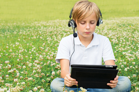 Pojke som sitter på en äng med blommande klöver med sin laptop i knät och hörlurar på sig.