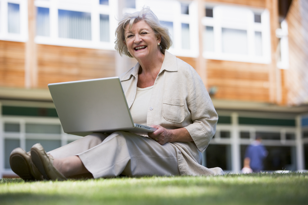 Äldre kvinna sitter i gräset utanför ett universitet med en laptop i knät.